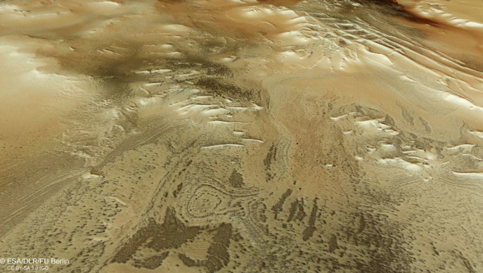 Esta imagen rectangular muestra parte de la superficie marciana como si el espectador estuviera mirando hacia abajo y a través del paisaje, con el suelo irregular y moteado apareciendo en tonos arrem