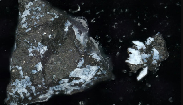 Imagen microscópica de una partícula oscura de Bennu, de aproximadamente un milímetro de largo, con una costra de fosfato brillante. A la derecha hay un fragmento más pequeño que se desprendió.