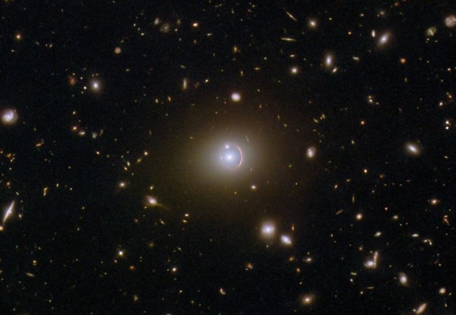 Esta imagen del telescopio espacial Hubble de la NASA/ESA muestra la galaxia remota HerS 020941.1+001557, que aparece como un arco rojo que rodea parcialmente una galaxia elíptica en primer plano.